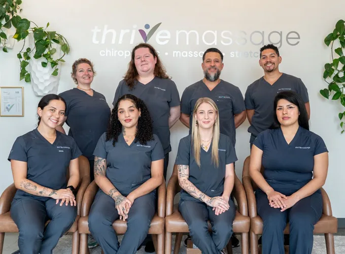 two rows of massage therapy staff sitting and standing in front of clinic name wall with greenery